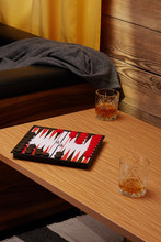 Staged Photo Of Backgammon On The Bedside Table With Whiskey Glasses Near The Couch And A Yellow Curtain. The Narde Set Consists Of A Folding Board, Black And Red Playing Chips And Two Cubes. 