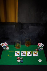 Staged photo of playing cards, casino chips and whiskey glasses on the black table against arm-chairs and a yellow curtain. The scene of the poker game is made in semi-darkness. 