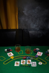 Staged photo of playing cards, casino chips and whiskey glasses on the black table against arm-chairs and a yellow curtain. The scene of the blackjack game is made in semi-darkness. 