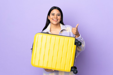 Wall Mural - Young latin woman woman isolated on purple background in vacation with travel suitcase and with thumb up