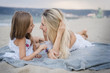 © Alla - Mom with daughters blondes in white dresses laugh, hug and lying on a gray knitted blanket near the blue sea on the beach at sunset.