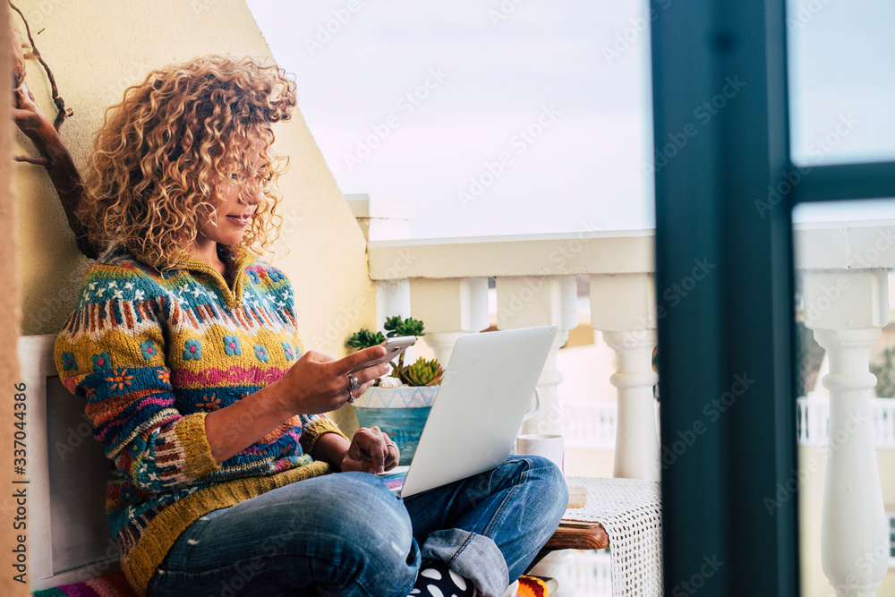 Beautiful adult caucasian blonde curly modern woman use phone device and personal laptop computer at home - concept of internet connection and roaming device - free office house smart work concept - obrazy, fototapety, plakaty 