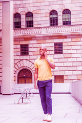 Wall Mural - Young African American Man wearing yellow short sleeve shirt, blue pants, white sneakers, carrying laptop computer, walking outside office in New York, talking on cell phone. Color filtered effect..