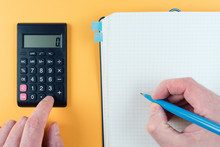 Close-up Shot Of Person Using Pocket Calculator While Taking Notes On Note Pad Against Orange Background