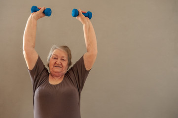  Health-conscious. Charming upbeat elderly lady working out with dumbbells and smiling pleasantly at the camera at home.