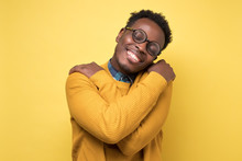 Pleased African American Man In Yellow Clothes And Glasses Hugs Himself, Has High Self Esteem. Studio Shot On Colored Wall.