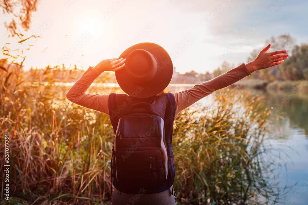 Traveler with backpack relaxing by spring river at sunset. Young woman raised arms feeling free and happy - obrazy, fototapety, plakaty 
