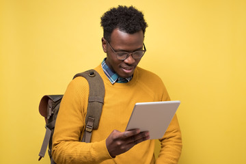 Sticker - Happy african american college student in glasses holding tablet on yellow wall. Studio shot