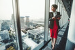 © skyNext - A good-looking African-American woman entrepreneur in a red skirt and black jacket is using a digital tablet while leaning against a panoramic window of a business office high-rise, cityscape outside