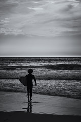 Wall Mural - Lone surfer walks into the ocean to catch a wave. View on an Atlantic coast and dramatic cloudy sky at sunset. Concept of outdoor activities. Lisbon, Portugal.High Contrast. B&W color.