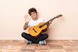 © luismolinero - Young caucasian man with a guitar sitting on the floor saluting with hand with happy expression
