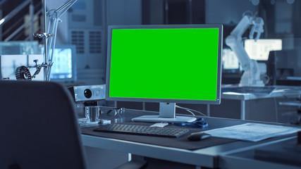 Poster -  On the Desk Computer with Isolated Green Mock-up Screen Display. In the Background Robot Arm Concept Standing in Heavy the Dark.Industry Engineering Facility
