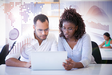 Wall Mural - Colleagues watching presentation and virtual statistic graphics. Business man and woman sitting at table in office, using laptop, looking at screen and talking. Working together concept