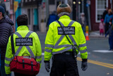 Fototapeta  - Two medical first responders walking in a street with people in the background. The officers are wearing a bright yellow reflective coat with grey stripes and a red first aid bag. 