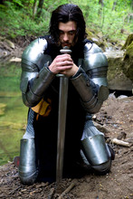Portrait Of Knight Leaning On Sword While Kneeling At Riverbank In Forest