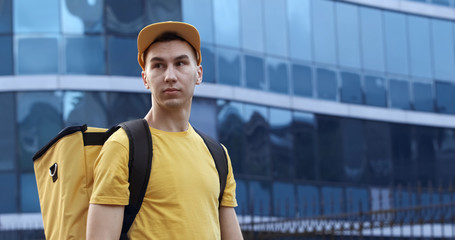 The courier in yellow uniform with a thermo backpack on the background of a business center with blue windows.