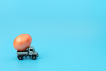 The Grey Truck Car Is Carrying An Orange Easter Egg On Blue Background. Happy Easter Holiday, Minimalism Concept. Greeting, Invitation Card With Copy Space.