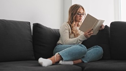 Sticker - A smiling young woman is reading a book while sitting on the couch in the morning