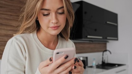 Canvas Print - A positive smiling young blonde woman is using her phone in the kitchen in the morning