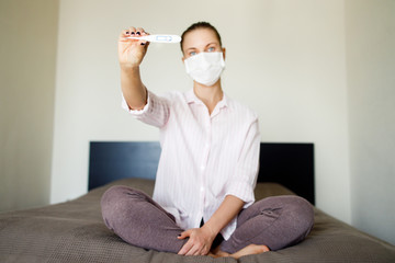 Female in medical mask looking at camera with thermometer in hand, with laptop . Coronavirus epidemic