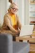 © AnnaStills - Senior woman in eyeglasses sitting on sofa and working online she typing on laptop computer at home