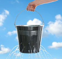 Woman Holding Leaky Bucket With Water Against Blue Sky, Closeup