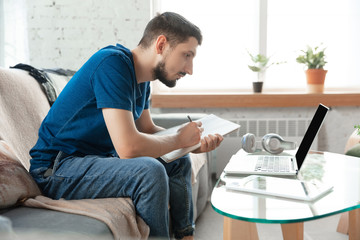 Young focused man studying at home during online courses or free information by hisself. Becomes driver, manager, photographer while isolated, quarantined. Using laptop, smartphone, headphones.