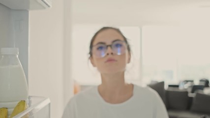 Wall Mural - Concentrated pretty brunette woman in eyeglasses looking for something in refrigerator on kitchen at home