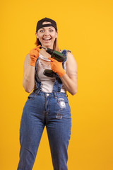 Builder girl in denim overalls and a black cap holds a drill and a screwdriver in her hands and poses on a yellow background