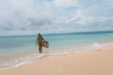 Beautiful and young girl in a dress posing on the shore of the ocean and sea with blue waves. Woman in a long dress model and fashion posing. Relax and relaxation on the island of Bali