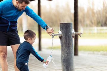 Funny little boy filling his water bottle in public fountain while his father helps him.