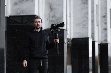 Wall Mural - Young Professional videographer holding professional camera on 3-axis gimbal stabilizer. Pro equipment helps to make high quality video without shaking. Cameraman wearing black hoodie making a videos.