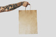 cropped photo on a hand with tattoos that holding blank craft paper package, bag mock-up. Empty space for design. White background