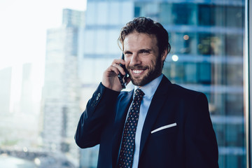 Wall Mural - Cheerful prosperous businessman in formal wear making cellular call standing near window in office interior, happy male executive manager satisfied with good news during mobile phone conversation