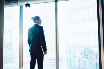 Wall Mural - Pensive caucasian male director of corporation standing near big windows in office interior pondering about future plans, back view of thoughtful businessman dressed in formal suit looking outside