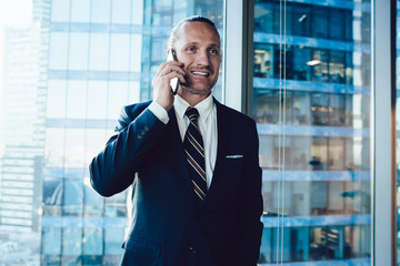 Wall Mural - Smiling prosperous businessman in formal outfit standing near office with mobile phone making call for discuss work, confident male entrepreneur dressed in elegant wear enjoying cellular talk