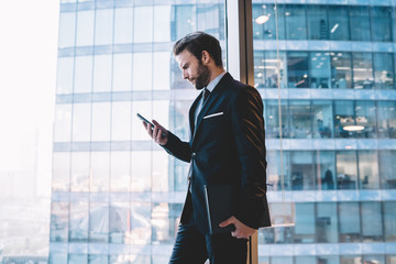 Wall Mural - Serious confident male manager in suit holding mobile phone checking notification about income mail near panoramic windows in office, confident businessman read information from web page on cellular