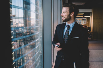 Wall Mural - Cheerful caucasian male entrepreneur in formal outfit holding mobile phone connected to 4G looking at window smiling, positive businessman satisfied with modern smartphone for communication and career