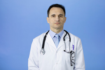 Wall Mural - Portrait of a caucasian adult doctor in a white coat with a stethoscope over neck on a blue background. Serious stern look at the camera. Glasses for the eyes are in the chest pocket. Studio shot