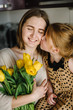 © Serhii - Cute little girl greeting mother and gives her a bouquet of flowers tulips on kitchen and kissing. Mother's day concept. Mom and daughter smiling. Happy family holiday and togetherness.