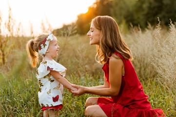 Young charming mother in a red dress crouched near her little daughter in the field..