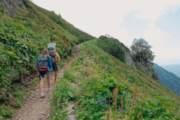 Young beautiful family, parents and child in the child carrier, hiking on the picturesque mountain trail in the summer forest with great mountain view with blue sky backwards, sunny day 