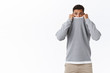 Scared, timid and cute young guy feeling afraid of horror movies, pulling sweater on face and frowning with arched sad expression, being frightened, standing shaking fear white background