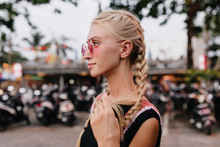 Pensive Blonde Woman In Black Attire Posing On Blur Street Background. Outdoor Shot Of Serious Tanned Lady With Braids Wears Pink Sunglasses.