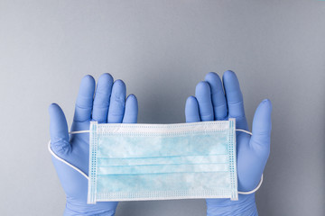 View of two hands with blue medical gloves holding a medical mask on a light gray background. Copy space design.