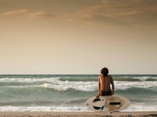 Rear View Of Shirtless Man Sitting On Surfboard At Beach