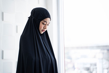 Wall Mural - portrait of beautiful asian muslim woman sitting closing her eyes meditating wearing black hijab rode covering the head, praying in a prayers private room with bright cool tone and white background