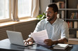 © fizkes - Happy millennial african american businessman in eyewear holding documents, doing paperwork, preparing report or analyzing market research results, working on computer in modern workplace office.