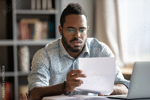 Fotografía Focused serious millennial african american businessman in glasses reading bank loan payment notification paper, sitting at table with computer