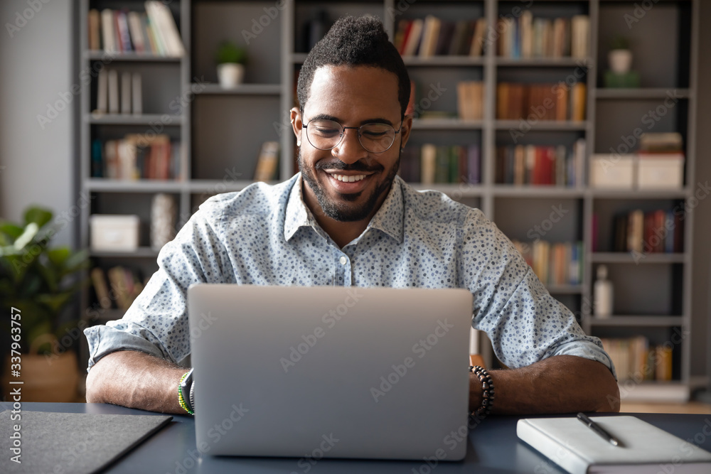 Front view head shot young smiling african american businessman in eyewear looking at laptop screen, reading pleasant messages in social networks, working on computer sitting at table in office home. - obrazy, fototapety, plakaty 